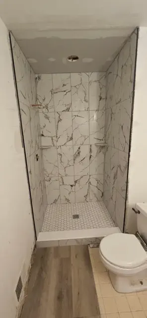 Narrow walk-in shower tiled floor to ceiling in marble-look tile with a small white hexagon floor