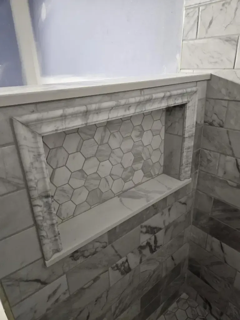 Shower niche lined with small marble hexagon mosaic, framed all round by a marble picture-frame trim in a marble-look wall