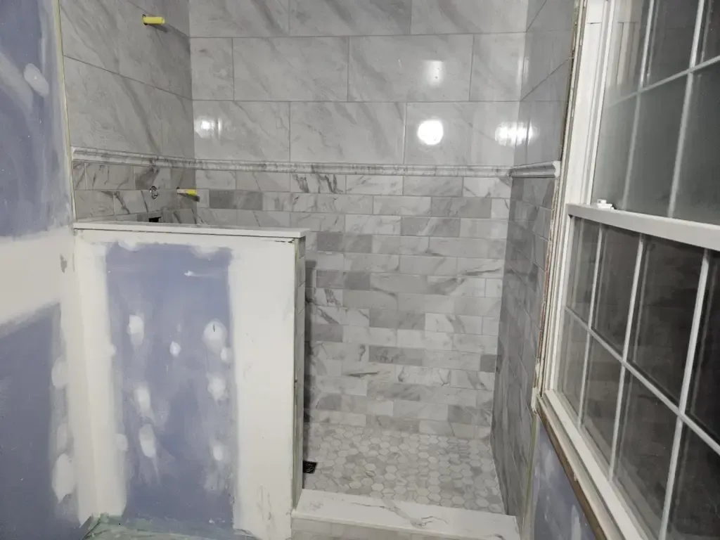 Marble-look shower beside a window, with a half wall and a hexagon floor, the drywall around it not yet painted